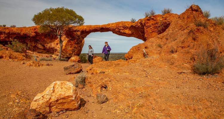 Twee bezoekers staan bij een natuurlijke zandstenen boog in het rode woestijnlandschap van Australië.