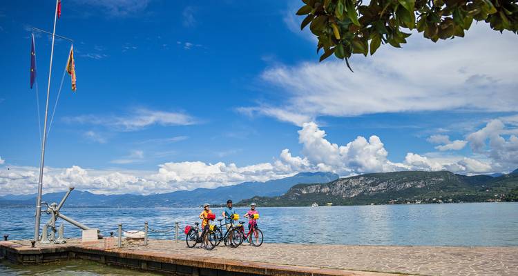 Trois cyclistes vêtus de maillots aux couleurs vives s'arrêtent à l'extrémité d'une jetée au bord d'un lac sous un ciel d'un bleu éclatant.