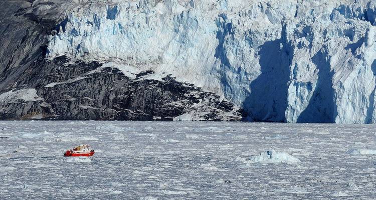 Un petit bateau de croisière navigue dans une mer de glace sous une paroi glaciaire imposante dans un fjord du Groenland.