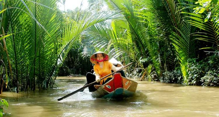 Une femme coiffée d'un chapeau conique pagaie dans une petite embarcation en bois le long d'un étroit canal tropical bordé de palmiers verts luxuriants.