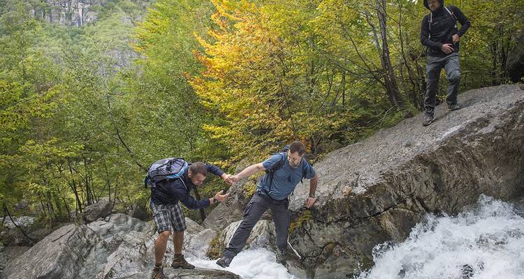 Tres excursionistas ayudándose mutuamente para cruzar un arroyo en un sendero rocoso.
