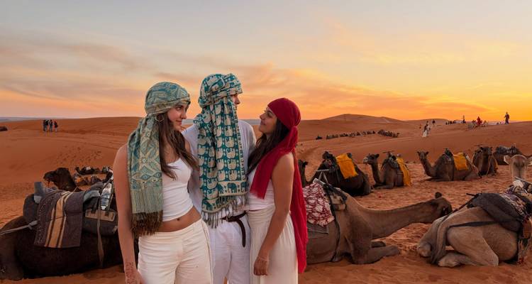 Groupe d'amis portant des foulards à motifs debout parmi des chameaux au repos sur fond d'un coucher de soleil vibrant du Sahara