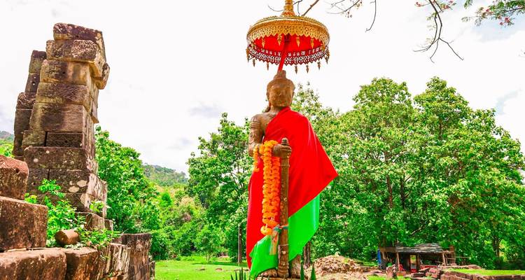 Statue de gardien en pierre drapée de tissu rouge et vert sous un parasol orné au milieu d'une forêt luxuriante au Wat Phou.