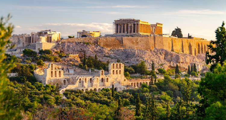 Iconic skyline of the Acropolis hill with the Parthenon glowing in golden evening light above green trees.