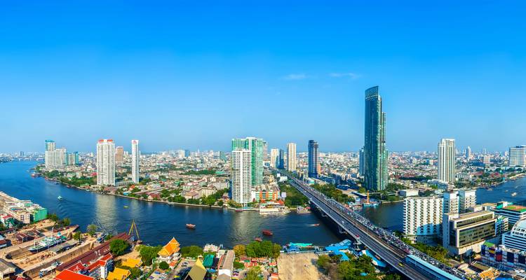 Wide aerial view of Bangkok’s skyline with the Chao Phraya River and tall modern skyscrapers under a bright sky.