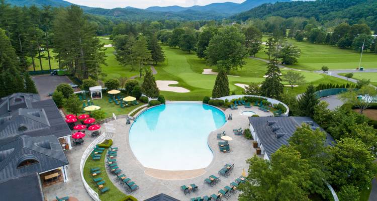 Aerial view of a luxurious resort pool surrounded by golf greens and distant forested mountains.