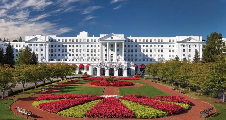 Grand white façade of The Greenbrier resort framed by manicured flower beds under a deep blue sky.