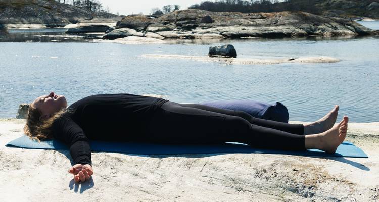 Una persona descansa en posición de savasana sobre una colchoneta de yoga encima de rocas calentadas por el sol junto a una bahía tranquila.