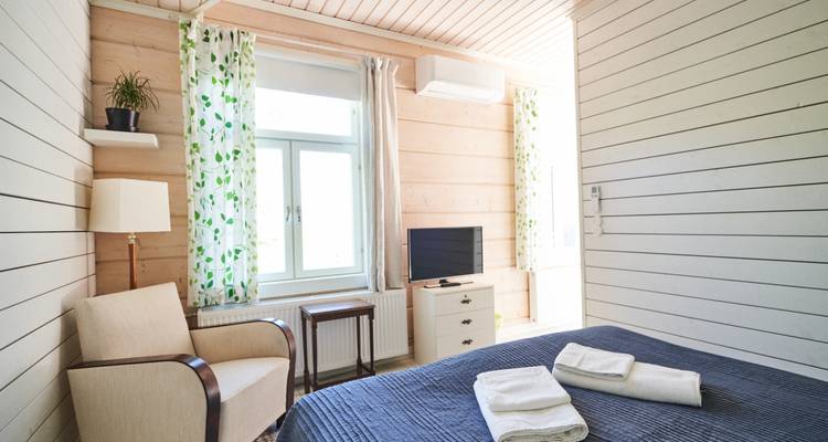 Habitación nórdica luminosa con paredes de paneles de madera, cortinas con motivos de hojas, toallas cuidadosamente dobladas sobre una colcha azul y un televisor pequeño.