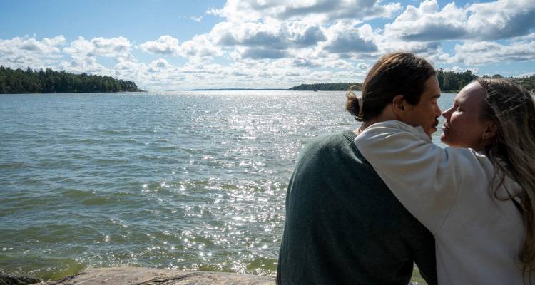 Una pareja joven comparte un momento tranquilo en una orilla rocosa con vista al mar brillante y la costa lejana llena de árboles.