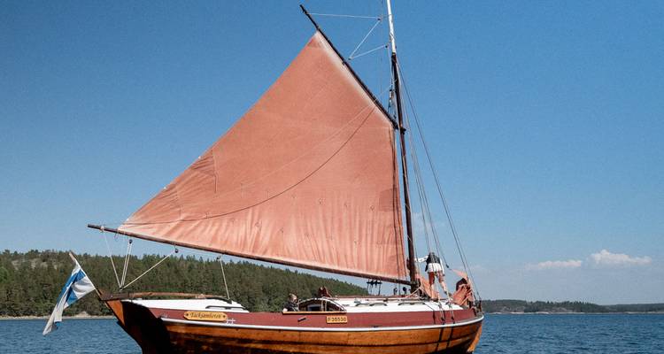 Un velero de madera tradicional con una vela triangular de color oxidado se desliza sobre las tranquilas aguas azules del Báltico.