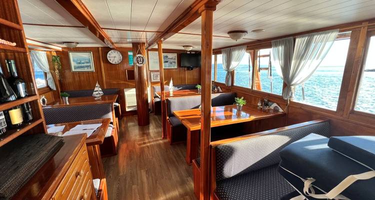 Área de comedor dentro de un barco con mesas de madera y ventanas con vista al mar.
