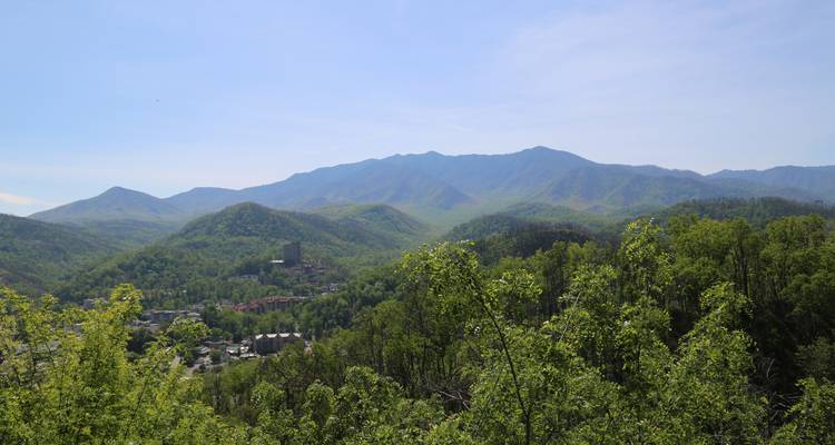 Green wooded hills surround Gatlinburg with distant Smoky Mountain peak under clear sky.