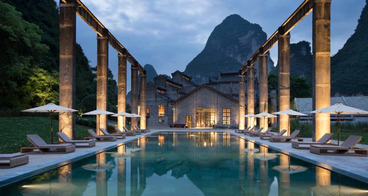 Illuminated industrial-chic pool courtyard framed by karst peaks at twilight.