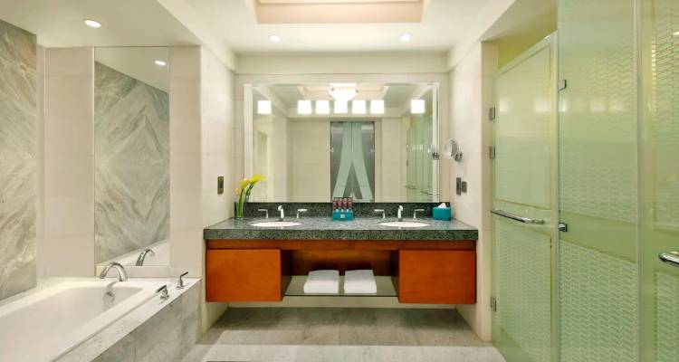 Modern, spacious hotel bathroom with marble surfaces, bathtub, dual sinks and large illuminated mirror.