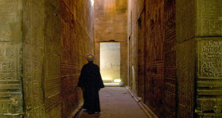 Dimly lit corridor of an ancient Egyptian temple with hieroglyphs and a lone person in silhouette.
