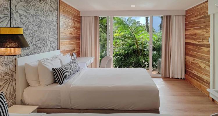 Chambre d'hôtel moderne et lumineuse avec papier peint tropical et grandes fenêtres donnant sur une végétation luxuriante.