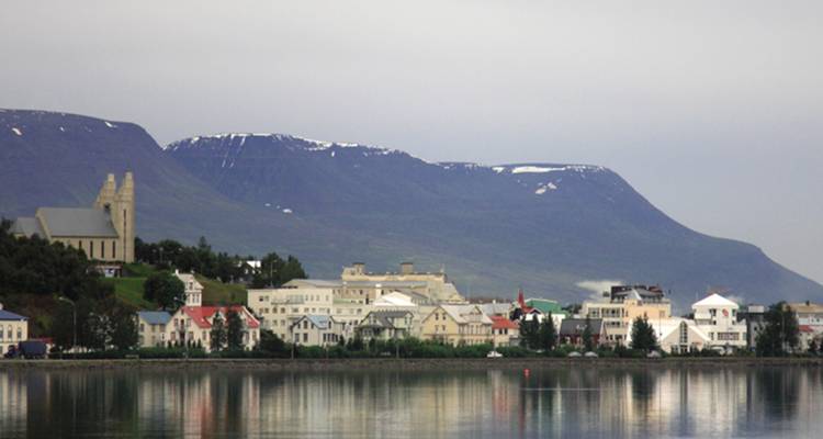 Pueblo junto al agua con iglesia y casas de colores vivos se sitúa bajo montañas con las cimas nevadas en un día nublado.