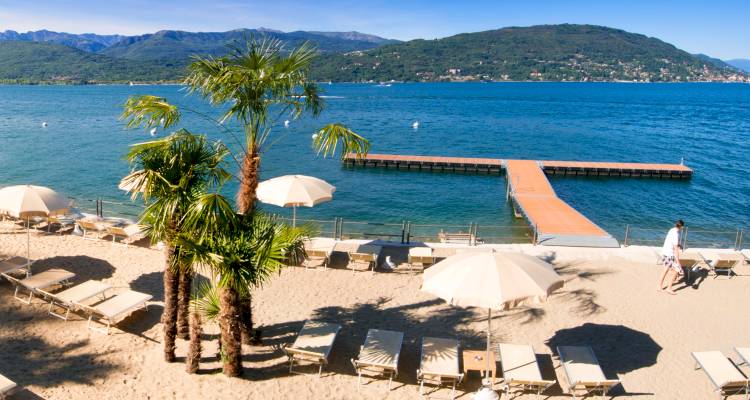 Playa privada arenosa con palmeras, sombrillas y tumbonas frente a un lago azul tranquilo y colinas verdes.