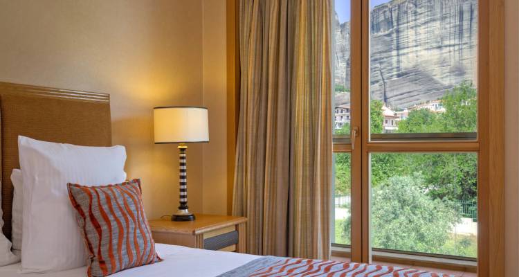 Close-up of hotel room corner with lamp, bed and towering Meteora cliffs visible outside window