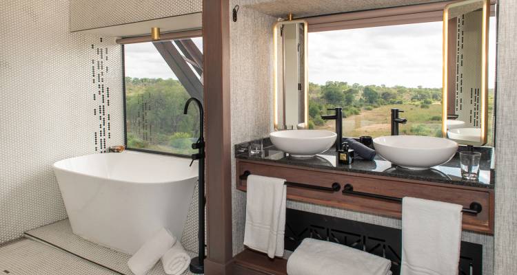 Modern safari lodge bathroom with freestanding tub and double sinks overlooking bushland through picture windows.