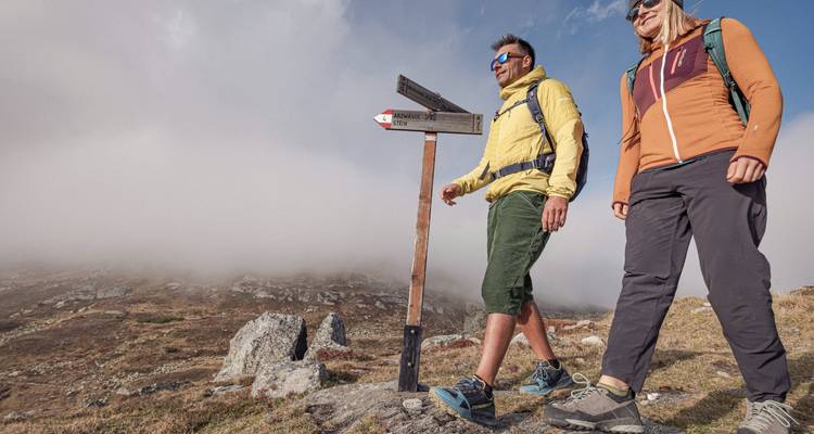 Excursionistas caminando junto a una señal en un sendero rocoso con montañas brumosas.