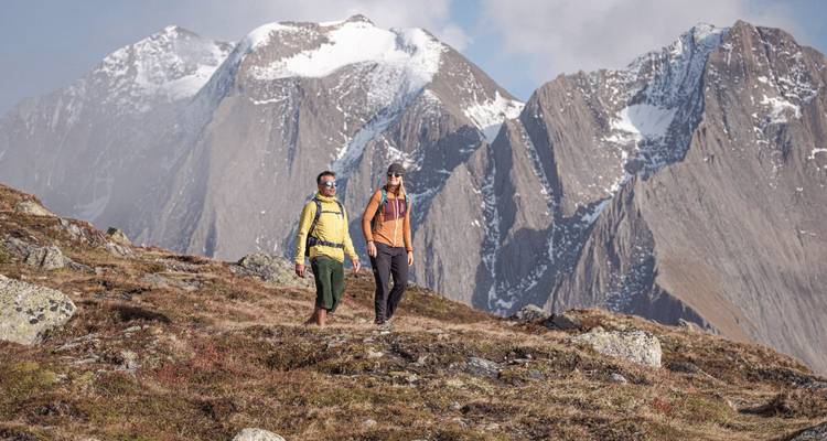 Dos excursionistas caminando por un sendero de montaña con picos nevados al fondo.