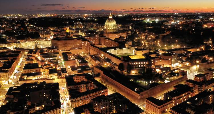 Vue aérienne nocturne de la Cité du Vatican et de Rome illuminées sous un ciel crépusculaire orange profond.