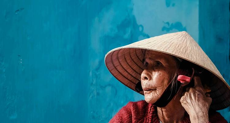 Femme vietnamienne âgée portant un chapeau conique qui regarde pensivement devant un mur bleu vif.