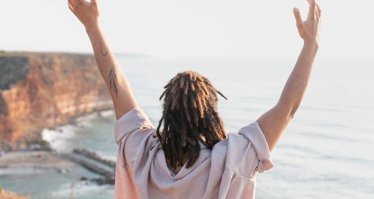 Back view of a traveler with raised arms overlooking ocean waves breaking on a cliff coast.