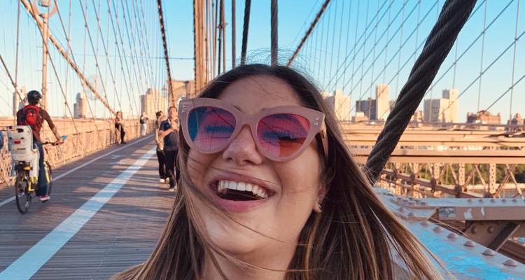 Smiling woman wearing oversized sunglasses takes a selfie on a suspension bridge at sunset light.