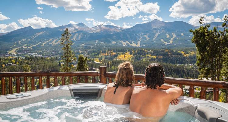 Couple relaxes in a steaming hot tub overlooking ski slopes and forested mountains.