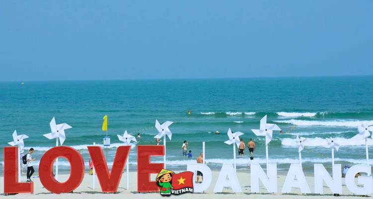 Una escena de playa con un letrero grande de 'LOVE DANANG' y gente junto al océano.