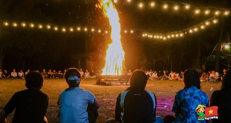 Un gran grupo se sienta alrededor de una fogata ardiente bajo cadenas de luces en la noche