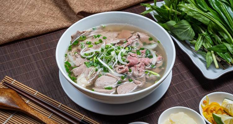 Tazón de pho con hierbas frescas y condimentos acompañantes sobre una estera de bambú.