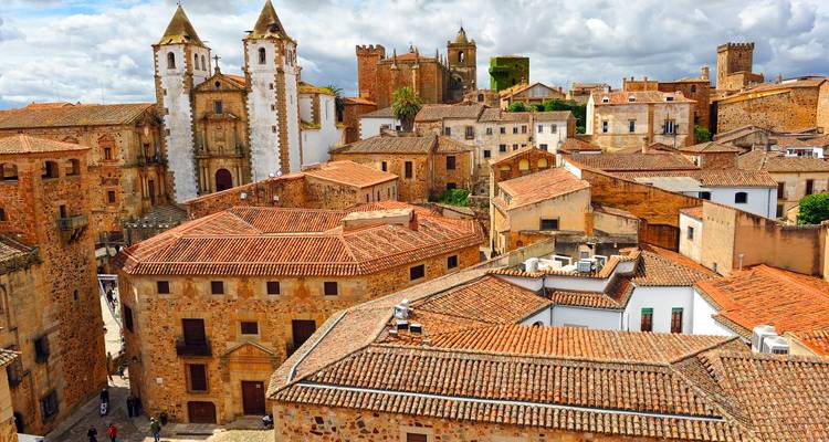 Historic rooftops and twin-towered church dominate the medieval stone townscape of Cáceres under patchy clouds.