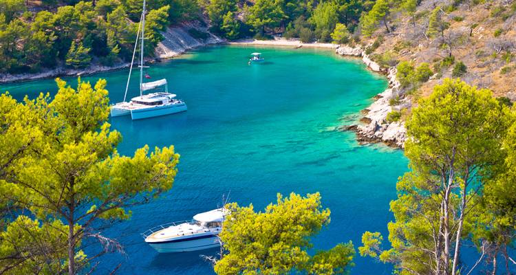 Secluded turquoise cove with sailing yachts surrounded by pine-clad cliffs.