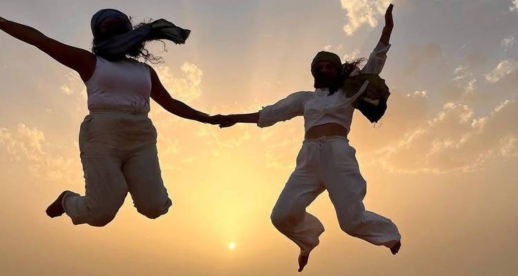 Two friends in headscarves leap joyfully against a glowing desert sunset sky.