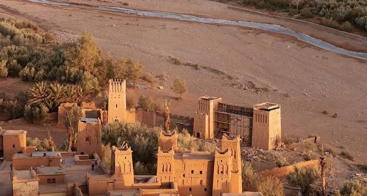 Aerial view of an ancient earthen kasbah complex beside a dry riverbed in the Moroccan desert.