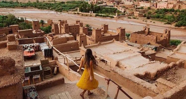 A traveler in a yellow dress gazes over the clay towers of an ancient kasbah village.