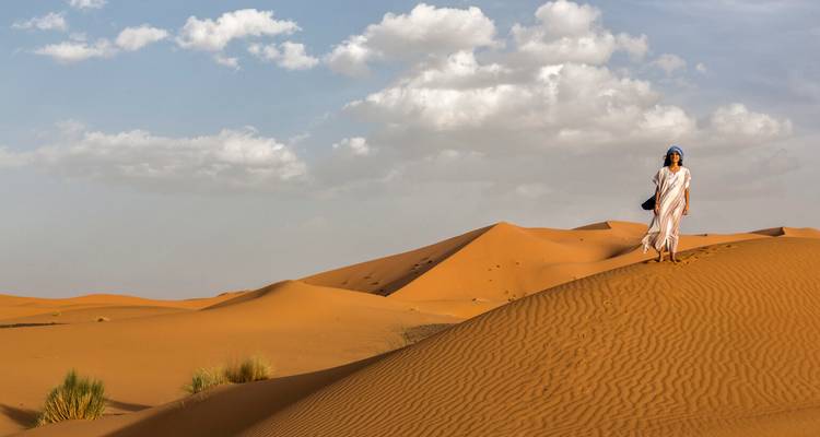 A lone traveler walks atop golden Saharan sand dunes beneath a bright sky.