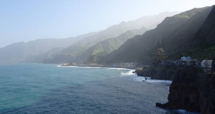 Côte escarpée de Santo Antão avec des crêtes vertes descendant vers une petite ville côtière et des vagues déferlantes.
