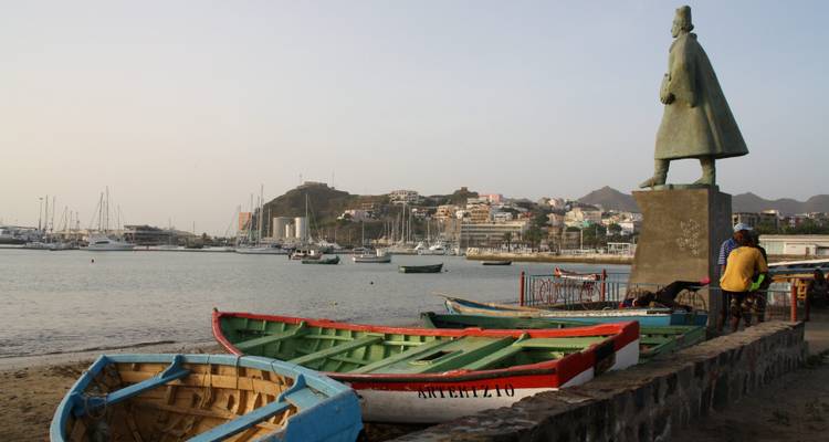 Barques de pêche au repos près d'une statue du front de mer et d'un port de plaisance avec la ville de Mindelo en arrière-plan.