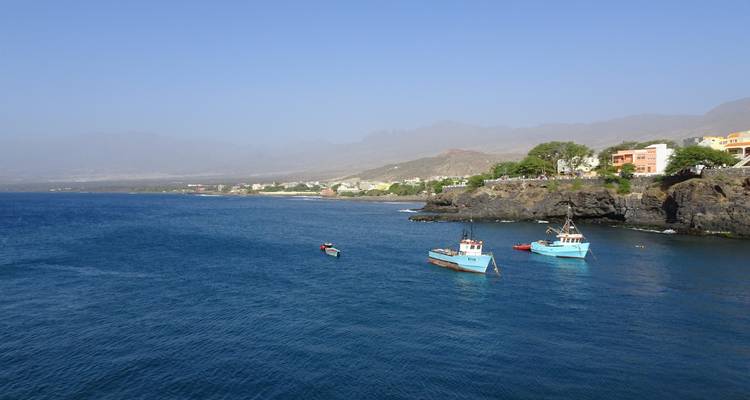 Baie bleue calme avec de petits bateaux de pêche ancrés près d'un promontoire rocheux et d'un village côtier.