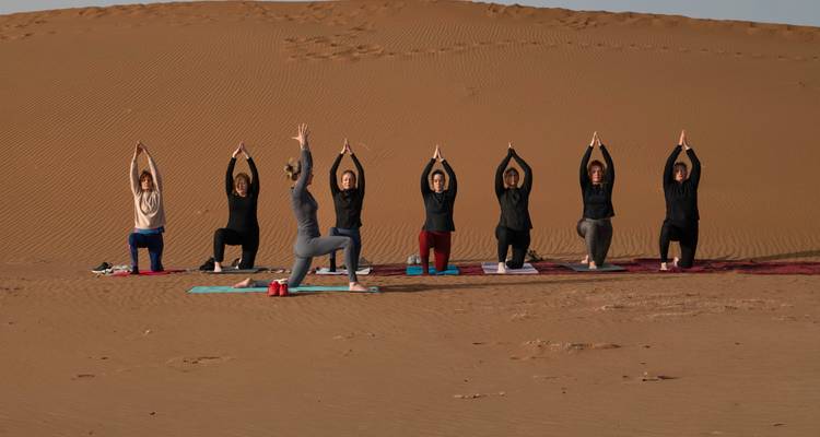 Series of yoga practitioners holding raised-arm poses on mats in front of a tall sand dune.