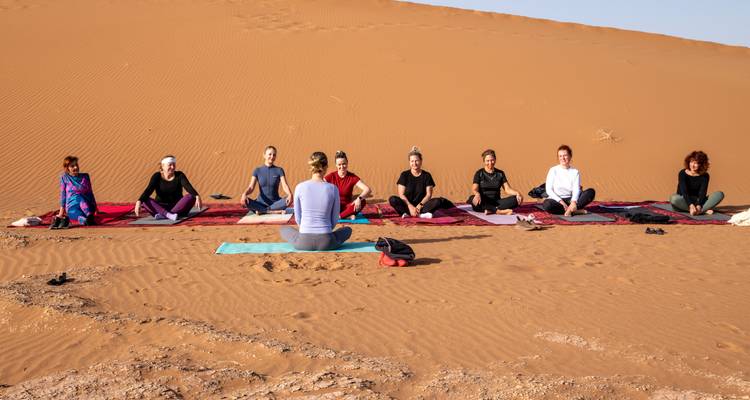 Yoga class seated cross-legged on rugs against the backdrop of a towering orange dune.