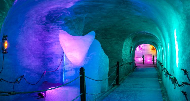 Un tunnel de glace éclairé au néon brille dans des nuances de bleu, violet et vert avec une passerelle à cordes menant vers l'avant.