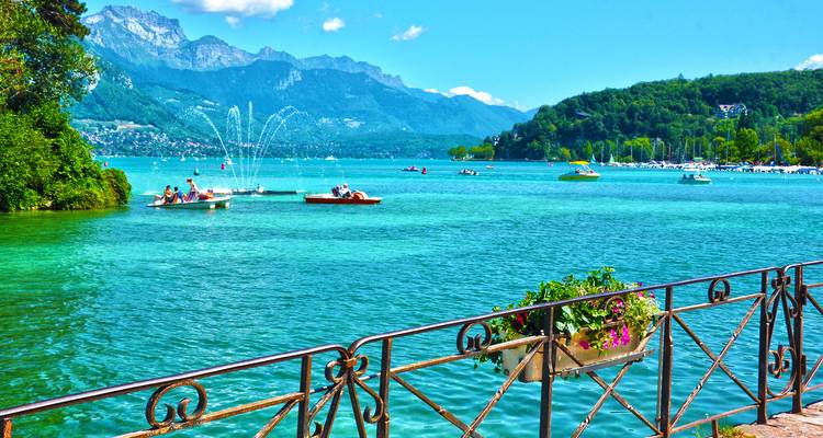 Des bateaux, des fontaines et des montagnes lointaines encadrent les eaux bleues scintillantes du lac d'Annecy par une journée ensoleillée.