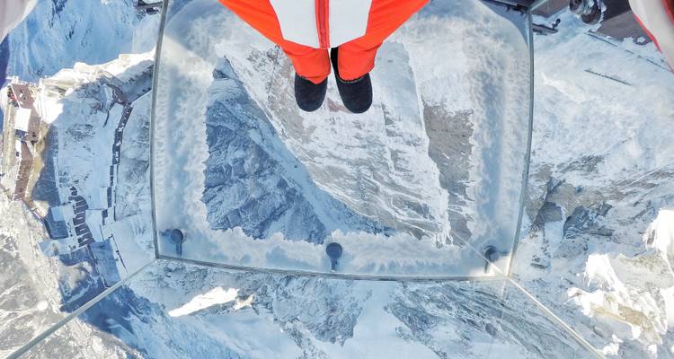 Un amateur de sensations fortes se tient sur un sol en verre regardant droit vers le bas sur des sommets enneigés depuis la plateforme 'Step into the Void'.