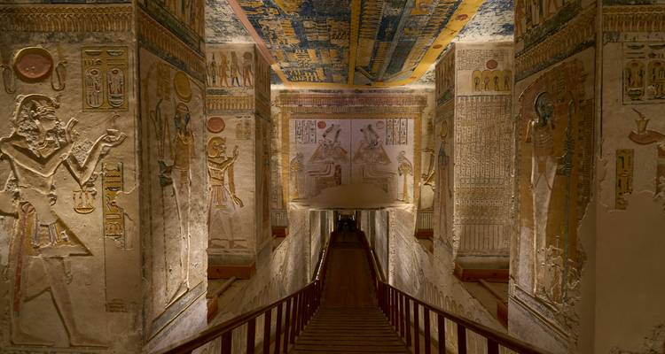 Decorated corridor inside an Egyptian tomb lined with painted reliefs and a wooden stairway.